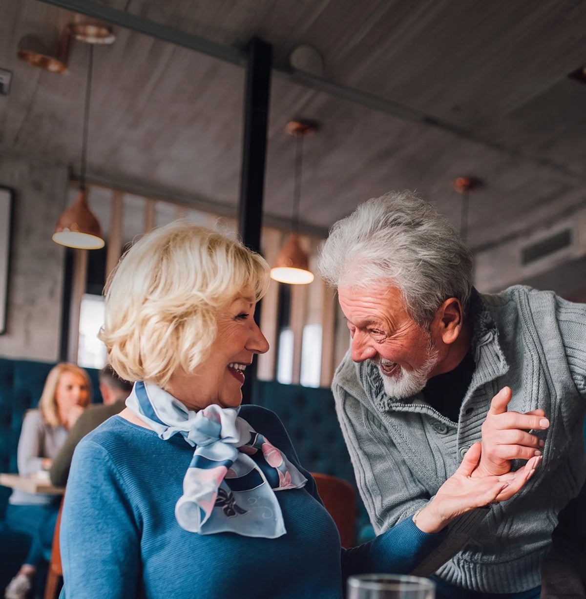 An older couple smiling and laughing in a cozy restaurant or cafe setting with warm pendant lights and a wooden ceiling.
