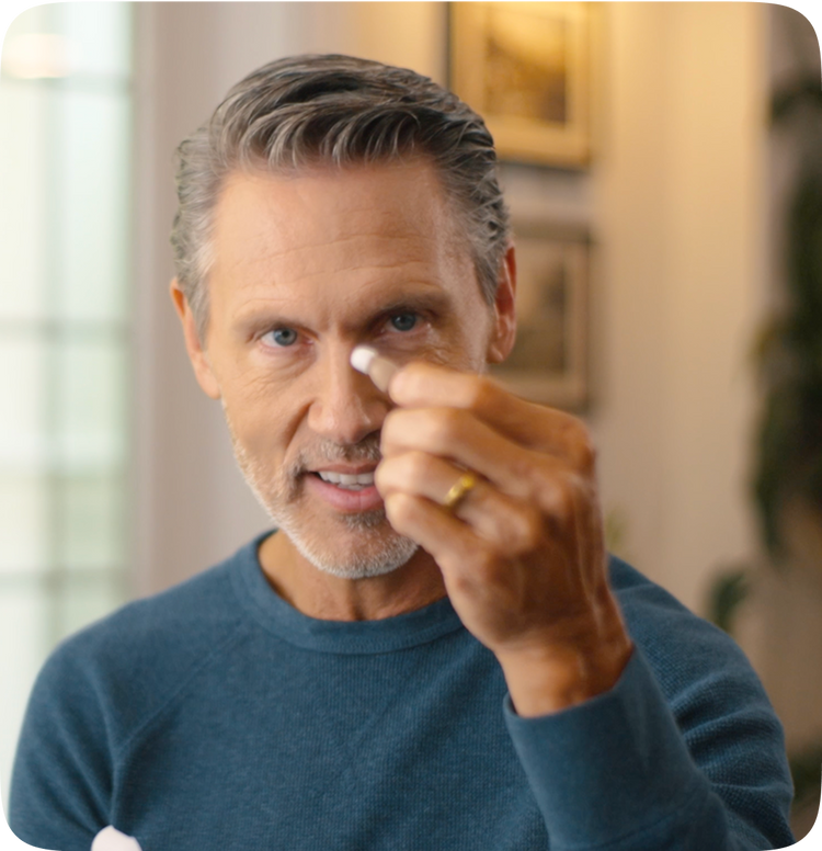 Middle-aged man in a blue sweater holding a small hearing aid between his fingers, smiling slightly while demonstrating it indoors.